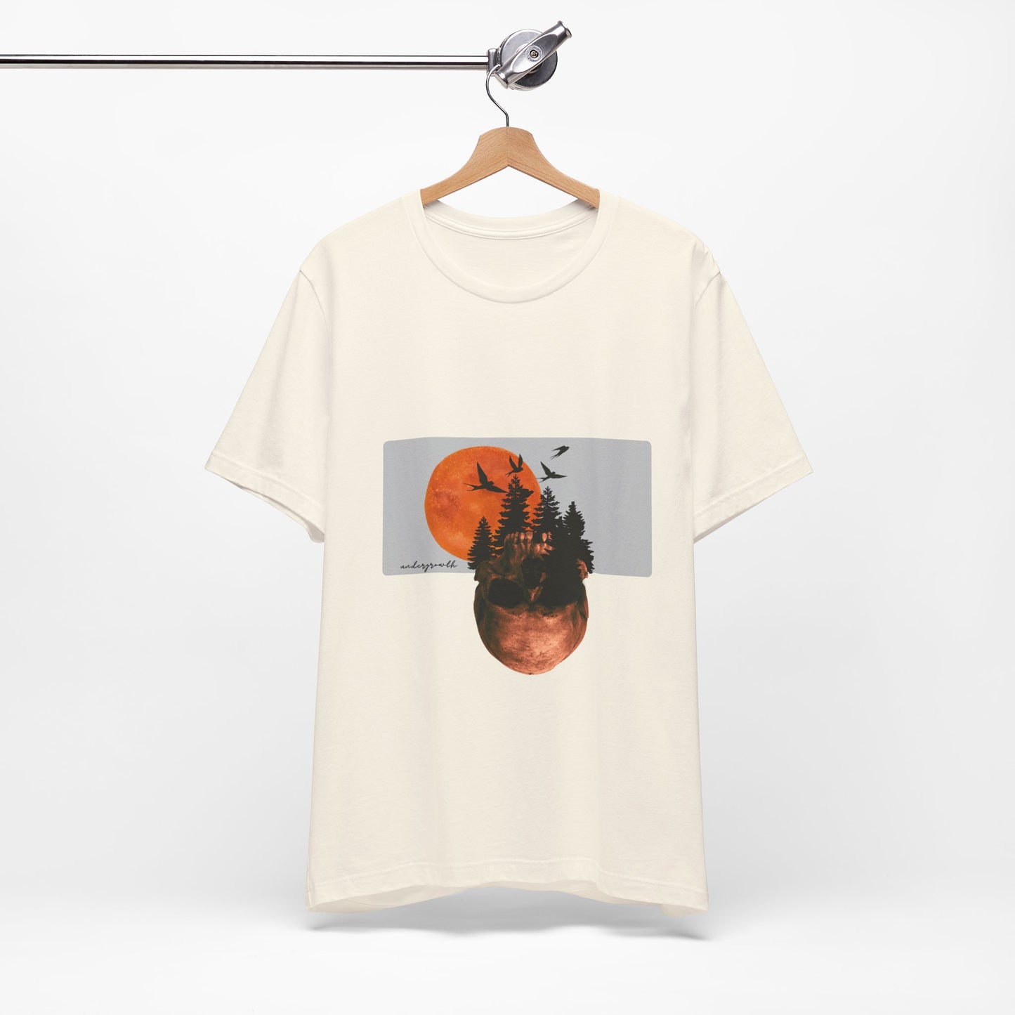 Beige t-shirt with a graphic design of trees and a sunset on a hanger against a white background